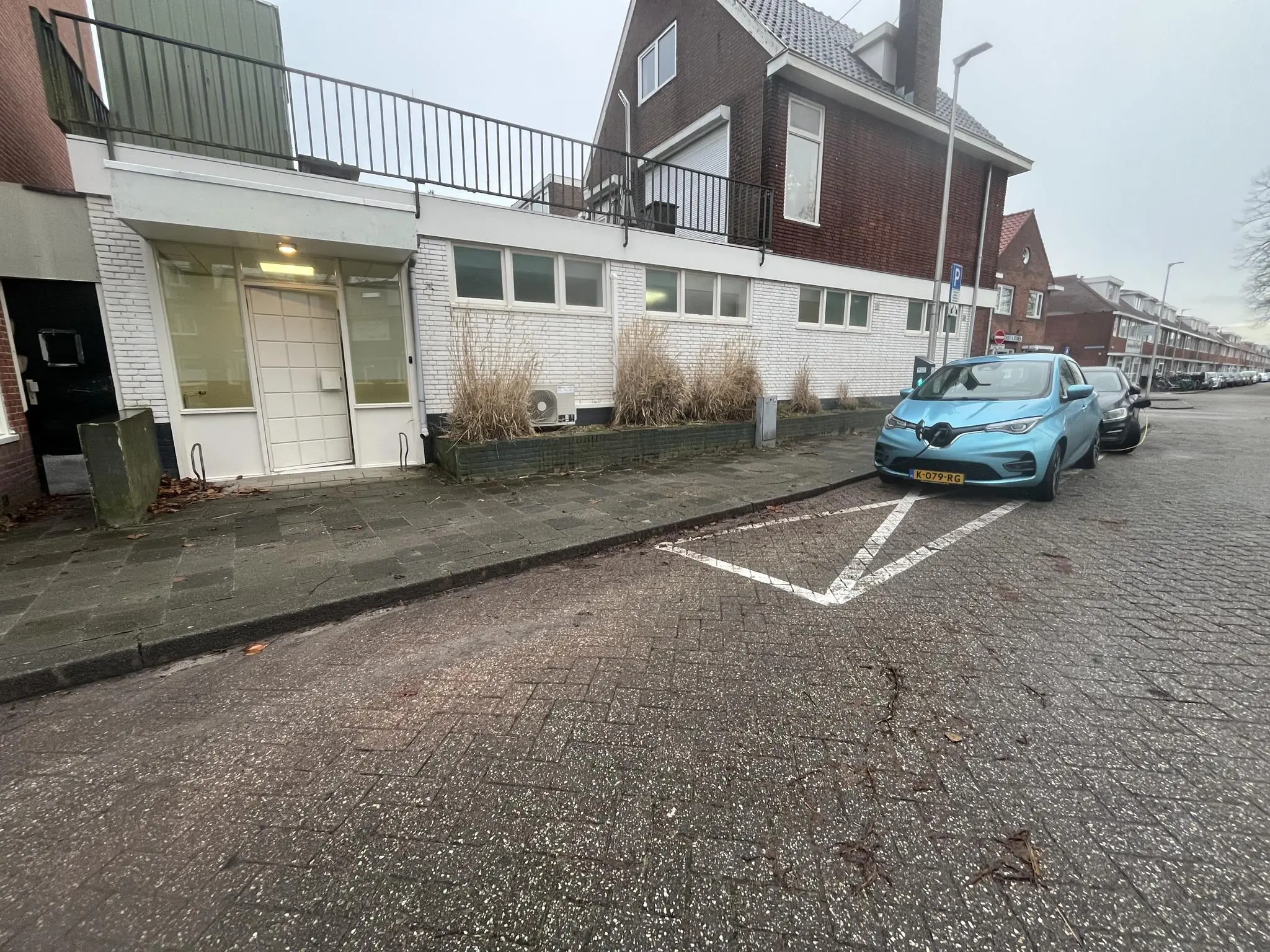 Een blauwe elektrische auto laadt op bij een laadpaal aan de Burgemeester Smitsstraat, naast een hoekpand met witte gevel en balkon.
